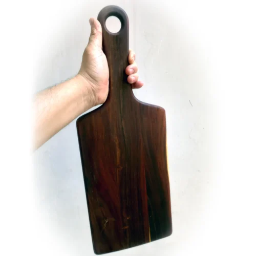 Hand holding rosewood paddle cutting board upright by the handle, showcasing full board height and color variation.